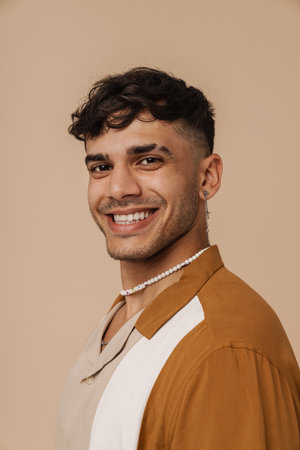 Brunette young man with earring smiling and looking at camera isolated over beige background
