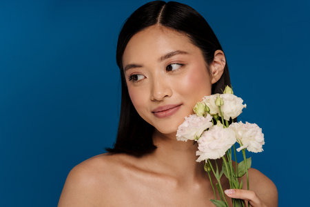 Portrait of young beautiful calm asian woman with white flowers looking aside, while standing over isolated blue backgroundの写真素材