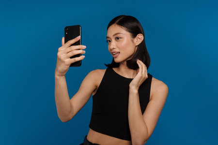Young beautiful smiling asian woman taking selfie on her phone and adjusting hair , while standing over isolated blue backgroundの写真素材