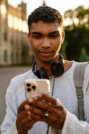 Young black man with headphones and backpack using mobile phone while walking by college outdoorsの写真素材