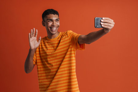 Young handsome smiling african man taking selfie on phone and waving , while standing over isolated...