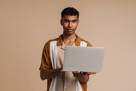 Young black man working with laptop while posing at camera isolated over beige backgroundの写真素材