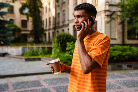 Young african man student in casual wear talking on cellphone and drinking coffee while standing outdoors in university campusの写真素材