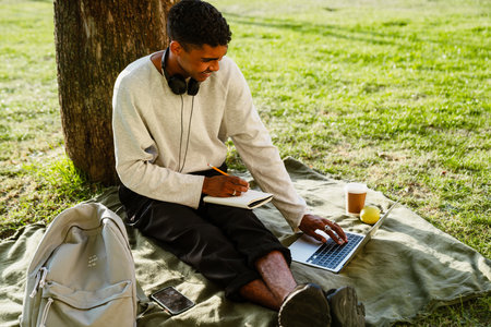Young black man studying with laptop and notepad while sitting under tree in parkの写真素材