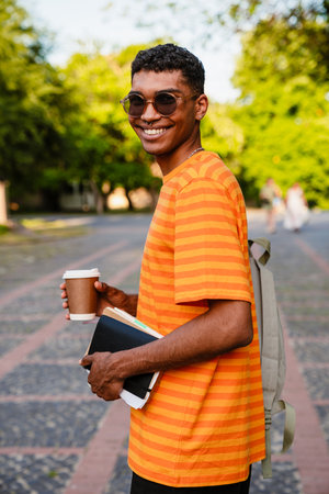 Young cheerful african man student in casual wear holding coffee and books while standing outdoorsの写真素材