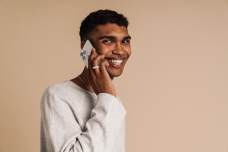 Portrait of young handsome stylish smiling african man talking phone and looking at camera , while standing with his side over isolated beige backgroundの写真素材