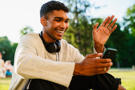 Young smiling african man in headphones having videocall and waving hand while sitting outdoors in parkの写真素材