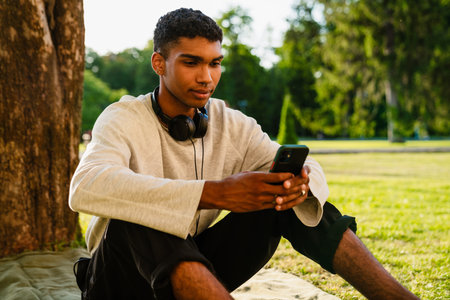 Young african man in headphones using mobile phone while sitting outdoors in parkの写真素材