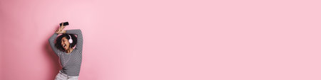 Cheerful young attractive african woman listening to music with headphones isolated over pink...