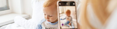 Mother taking photo of her blonde baby son on mobile phone at home