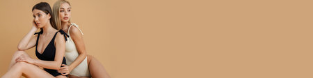 Two sensual elegant women in swimsuits posing together isolated over beige background