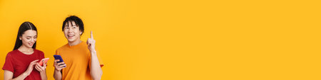 Young multiracial couple pointing finger upward and using cellphones isolated over yellow wallの写真素材