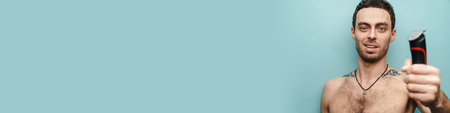 Close up portrait of a smiling middle aged shirtless man holding electric shaving over blue wall...