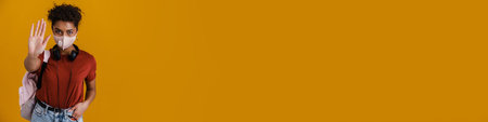 Young black woman in face mask showing stop gesture isolated over yellow backgroundの写真素材