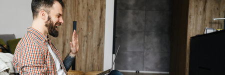 Happy mid aged brunette bearded man on a video call via laptop computer while sitting on a floor in the living roomの写真素材