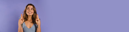 Young smiling woman holding fingers crossed for good luck isolated over purple backgroundの写真素材