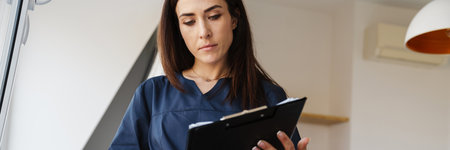 The brunette woman in blue medical uniform reading paper on the tablet while standing by the window in a bright roomの写真素材