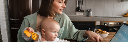 Smiling mid aged woman working on laptop computer while holding her baby on lap in the kitchen