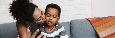 Black smiling woman taking selfie photo on cellphone while hugging her son at home