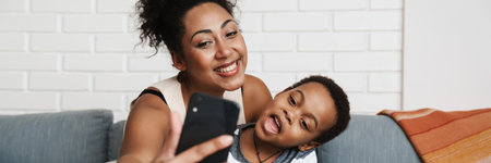 Black smiling woman taking selfie photo on cellphone while hugging her son at home
