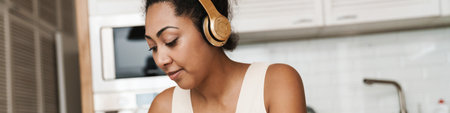 Black woman in headphones working with laptop while sitting at table in home kitchenの写真素材