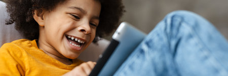Black curly boy smiling and using tablet computer while sitting on sofa at home