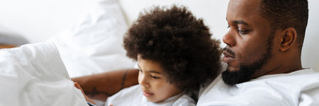Black father and son using tablet computer while lying in bed at homeの写真素材