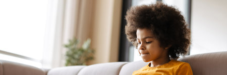 Black curly boy using tablet computer while sitting on sofa at homeの写真素材