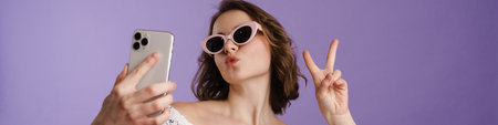 Happy young white woman holding mobile phone standing over violet background, taking a selfie,...