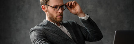 The concentrating man in a jacket and shirt looking into the laptop screen while straightening his glasses in the studioの写真素材