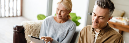Smiling middle aged white couple relaxing on a couch at home, relaxing, using tablet, writing in...