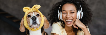 Young happy african woman in casual wear with beagle puppy at home, relaxing on bed in headphones laughingの写真素材