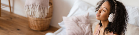 Smiling mid aged african woman listening to music with headphones while relaxing on bed at home, meditatingの写真素材