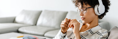 Young curly woman in headphones using laptop while sitting on armchair at home