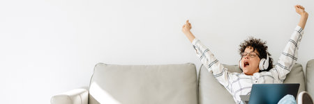 Young woman in headphones yawning and using laptop while sitting on couch at homeの写真素材
