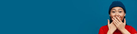 Happy young asian woman in casual clothes standing over blue wall background, covers mouthの写真素材
