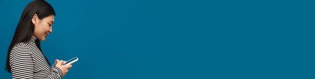 Brunette asian teen girl thinking while using mobile phone isolated over blue background