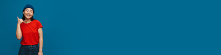 Asian teen girl in hat smiling and showing handset gesture isolated over blue backgroundの写真素材