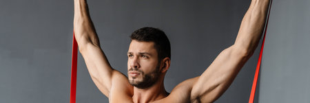 Young shirtless sportsman doing exercise with expander stretch tape indoors