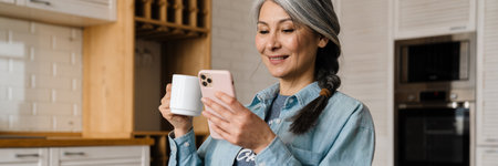 Mature grey woman smiling and using cellphone while drinking coffee at home