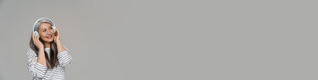 Mature white-haired woman smiling while listening music with headphones isolated over gray wallの写真素材