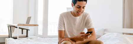 Smiling young hispanic man using mobile phone while sitting on bed at homeの写真素材