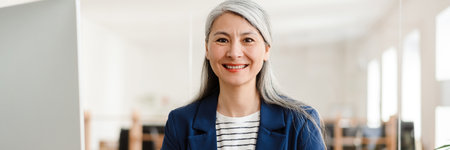 The smiling Asian woman sitting in the blue suit at the table and looking to the camera in the officeの写真素材