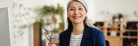The smiling Asian woman holding a tablet with papers and glasses in her hands and looking at the camera in a bright officeの写真素材
