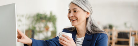 The smiling Asian woman with a cup in her hands sitting at the table and showing her finger to the computer monitor in a bright officeの写真素材