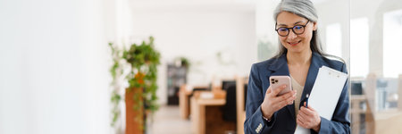 The smiling Asian woman in glasses and a suit looking at the phone while holding a tablet with papers in hands in a light officeの写真素材
