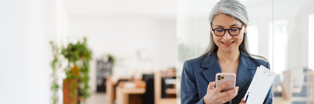 The pensive Asian woman in glasses and a suit looking at the phone while holding a tablet with papers in hands in a light officeの写真素材