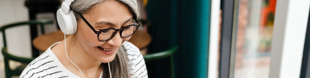 Mature smiling woman listening music with headphones and cellphone in cafe indoors