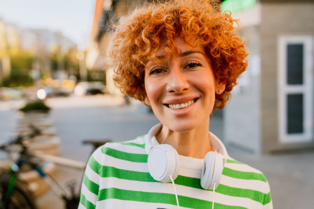 Ginger middle-aged woman standing outdoors with headphones over her neck and smiling at cameraの写真素材