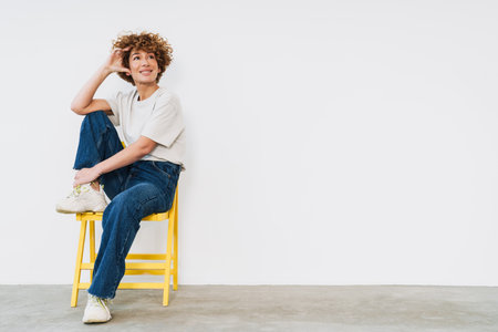 Middle-aged ginger woman sitting on yellow chair and looking away at copy space isolated over white...